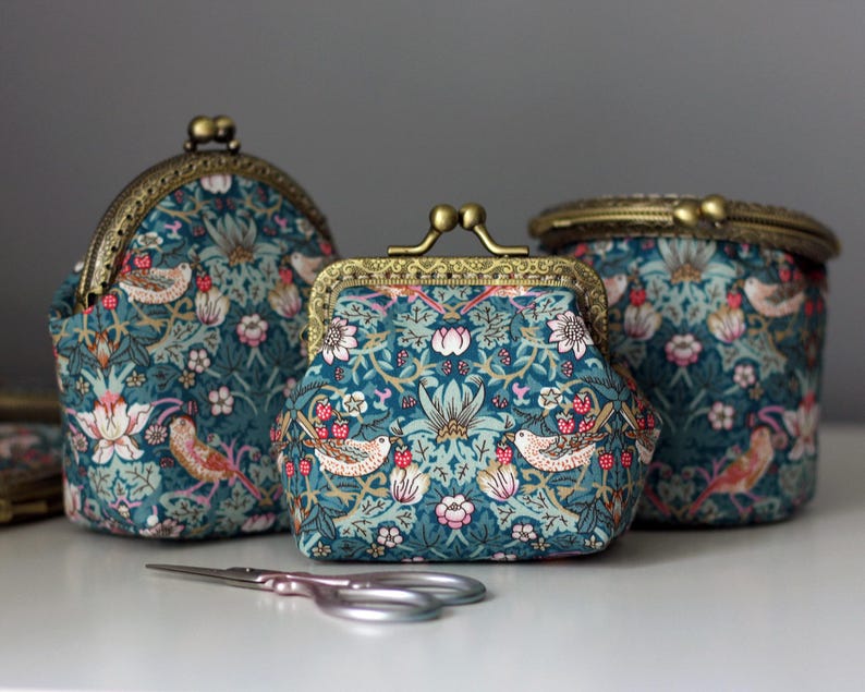 May include: Three teal coin purses with brass clasps. The purses feature a floral and bird pattern in shades of pink, white, and gold. A pair of small silver scissors rests in front of the purses.