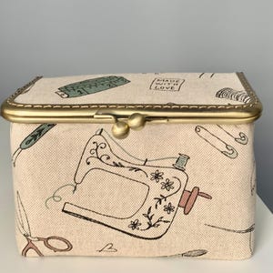 May include: A rectangular fabric storage box with a beige background featuring a black line-art pattern of sewing machines, scissors, safety pins, and measuring tapes. The box has a brass-colored metal frame with a dual-ball clasp closure.