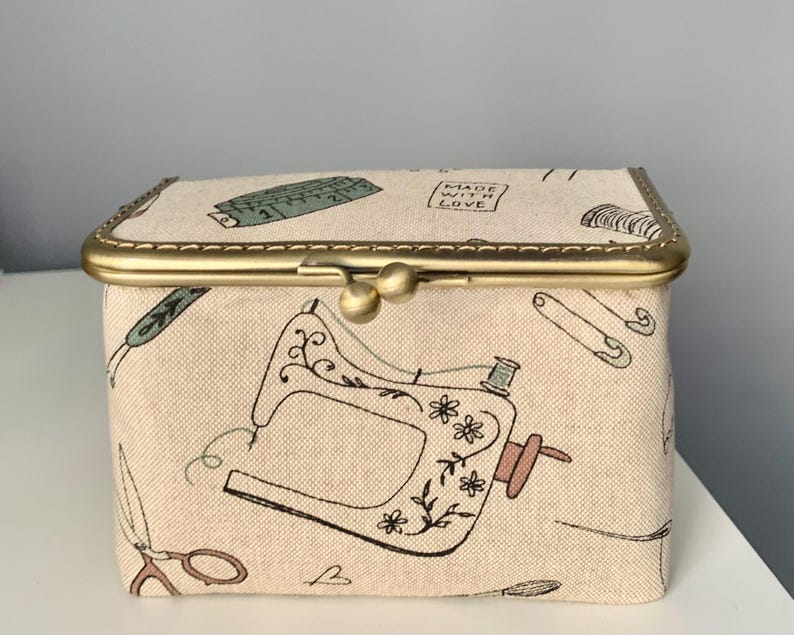 May include: A rectangular fabric storage box with a beige background featuring a black line-art pattern of sewing machines, scissors, safety pins, and measuring tapes. The box has a brass-colored metal frame with a dual-ball clasp closure.