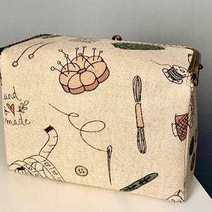 May include: A rectangular beige fabric box with a metal clasp, featuring a printed pattern of sewing supplies including a pincushion, measuring tape, buttons, needles, and thread. The text "hand made" with a small heart is printed on the side.