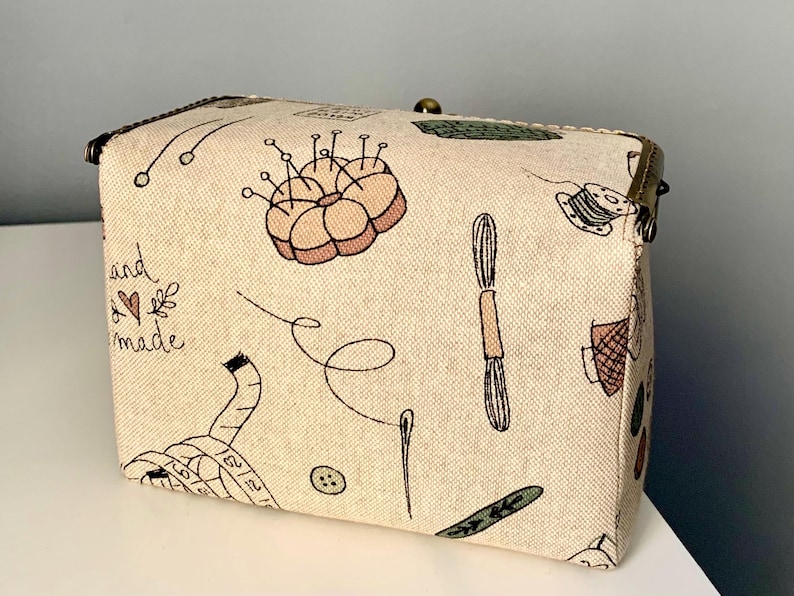 May include: A rectangular beige fabric box with a metal clasp, featuring a printed pattern of sewing supplies including a pincushion, measuring tape, buttons, needles, and thread. The text "hand made" with a small heart is printed on the side.