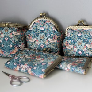 May include: A collection of five fabric pouches and purses featuring a teal floral and bird pattern with antique brass-toned metal kiss-lock clasps. The set includes various shapes and sizes, accompanied by a small pair of metal scissors.