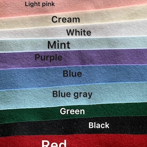 May include: A collection of felt fabric sheets arranged in horizontal layers, featuring colors labeled as Pink, Light pink, Cream, White, Mint, Purple, Blue, Blue gray, Green, Black, Red, Orange, and Yellow. Each strip displays a soft, matte texture.