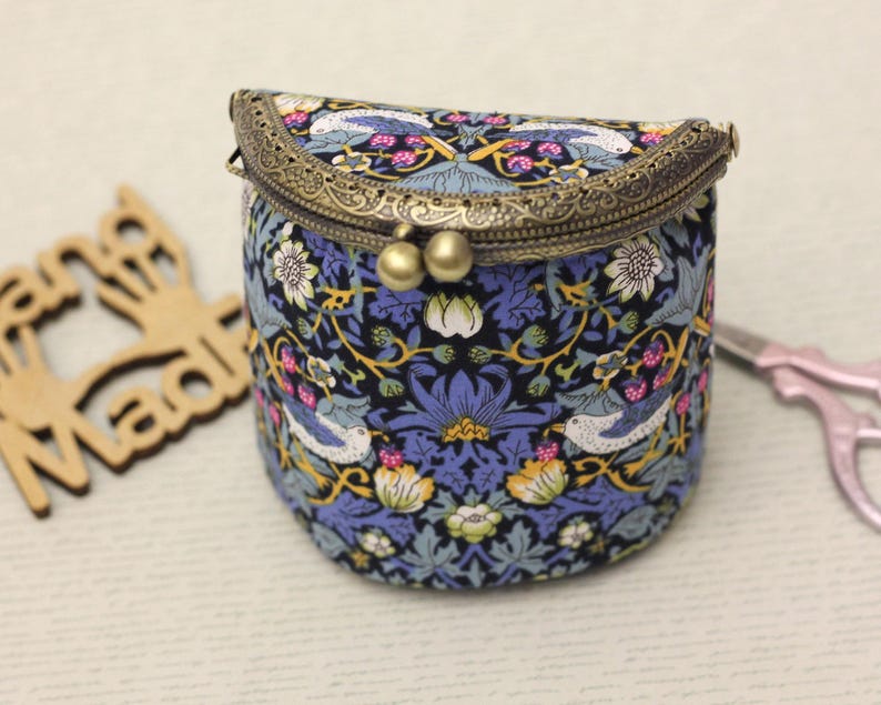 May include: A small, round, fabric purse with a decorative metal clasp. The purse features a blue and black floral pattern with birds and flowers. The clasp is a bronze color, and the purse has a vintage aesthetic.