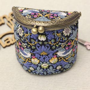 May include: A small, cylindrical coin purse with a decorative floral and bird pattern in blue, white, and yellow. The purse has a bronze-colored clasp and a hinged top. The background includes a wooden cutout and a pair of scissors.