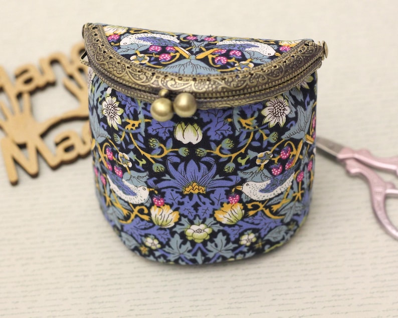 May include: A small, cylindrical coin purse with a decorative floral and bird pattern in blue, white, and yellow. The purse has a bronze-colored clasp and a hinged top. The background includes a wooden cutout and a pair of scissors.