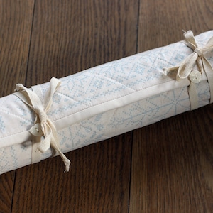 May include: A white fabric roll with a blue and white geometric pattern is tied with two white ribbons and buttons.