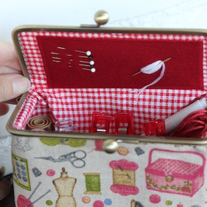 May include: A fabric sewing kit case with a metal kiss-lock frame. The exterior features a beige print of sewing supplies. The interior is lined with red and white gingham fabric and includes a red felt panel holding sewing pins and a needle.