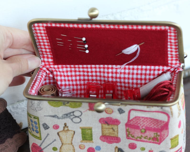 May include: A fabric sewing kit case with a metal kiss-lock frame. The exterior features a beige print of sewing supplies. The interior is lined with red and white gingham fabric and includes a red felt panel holding sewing pins and a needle.