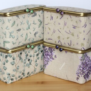 May include: Four rectangular fabric storage boxes with brass-toned metal frames and clasp closures. Two boxes feature green leaf patterns with green bead clasps, while two feature purple lavender floral prints with dark purple bead clasps.