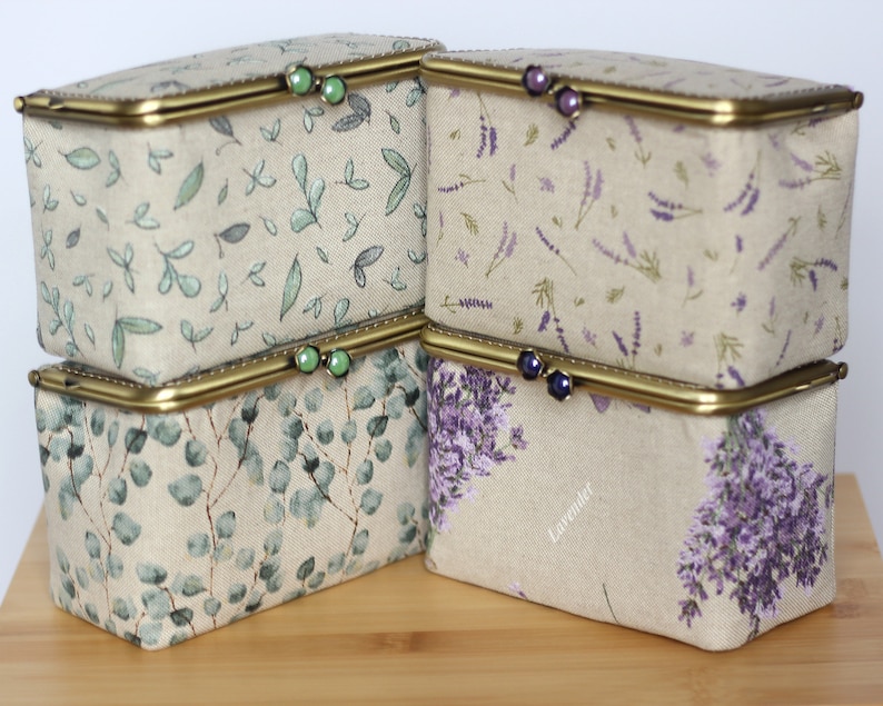 May include: Four rectangular fabric storage boxes with brass-toned metal frames and clasp closures. Two boxes feature green leaf patterns with green bead clasps, while two feature purple lavender floral prints with dark purple bead clasps.