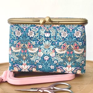 May include: A rectangular fabric pouch with a brass kiss-lock closure. The textile features a repeating pattern of birds, strawberries, and floral motifs in shades of teal, pink, and white. A pink plastic embroidery hoop and metal scissors are visible.