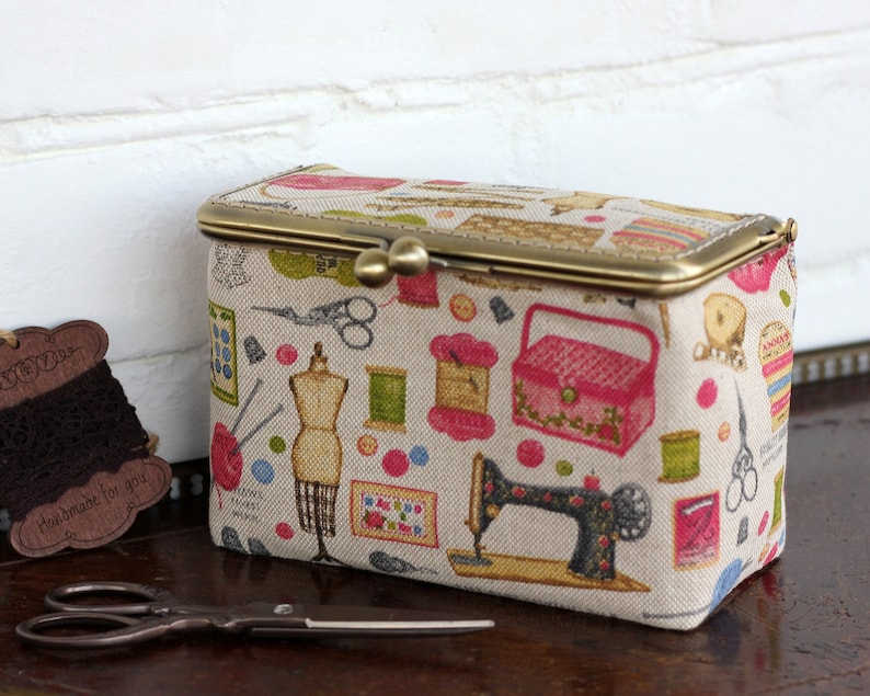 May include: A rectangular fabric storage box with a brass kiss-lock closure, featuring a beige background printed with colorful sewing-themed illustrations including a sewing machine, dress form, scissors, spools of thread, and a pink sewing box.