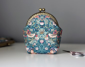 May include: A fabric coin purse with a teal background featuring a repeating pattern of birds, flowers, and berries. The purse has a metallic bronze-toned kiss-lock clasp at the top and a structured, rounded shape with a flat base.