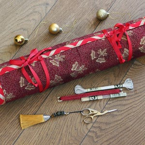 Può includere: Un rotolo di tessuto bordeaux con un motivo a campane dorate, legato con nastri rossi. Ornamenti natalizi dorati e forniture per ricamo, tra cui filo e piccole forbici dorate, sono disposti nelle vicinanze.