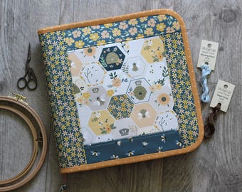 Puede incluir: Una carpeta cuadrada forrada en tela con un panel central de patrón hexagonal de abejas, colmenas y flores en amarillo, blanco y azul oscuro. El borde es un estampado floral a juego con un ribete de color amarillo mostaza.