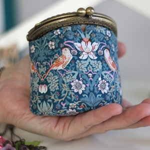 May include: A small, teal fabric coin purse with a brass clasp. The purse features a floral and bird pattern in shades of teal, orange, pink, and white. The clasp is a vintage style with two small, round closures.