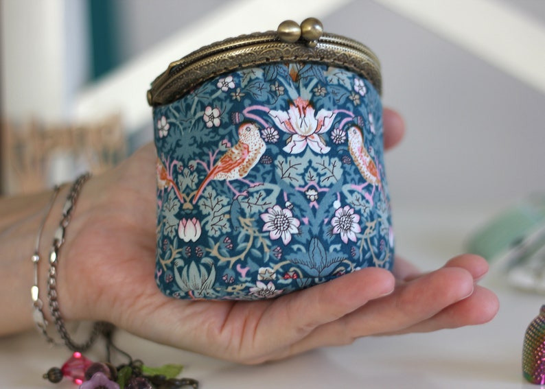 May include: A small, teal fabric coin purse with a brass clasp. The purse features a floral and bird pattern in shades of teal, orange, pink, and white. The clasp is a vintage style with two small, round closures.