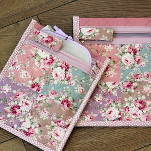 Może przedstawiać: Dwie pikowane płócienne saszetki z kwiatowym wzorem róż w kolorze różowym, lawendowym i turkusowym. Jedna saszetka ma zamek błyskawiczny i małe pasujące akcesorium. Druga saszetka ma zamek błyskawiczny i małe pasujące akcesorium.