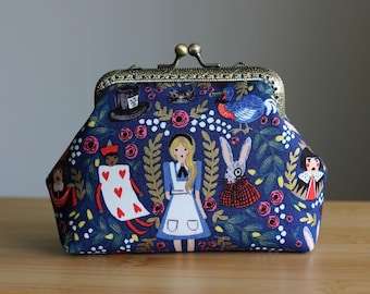 Puede incluir: Un monedero de tela azul marino con un cierre metálico de boquilla. El exterior presenta un estampado colorido con personajes que incluyen una niña, un conejo, una carta de juego y un sombrero de copa, rodeados de motivos florales y hojas.