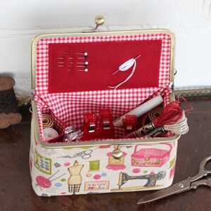 May include: A rectangular sewing organizer with a brass frame and clasp, featuring a beige fabric exterior with colorful sewing-themed illustrations. The interior is lined with red and white gingham fabric and contains sewing needles, thread, and clips.