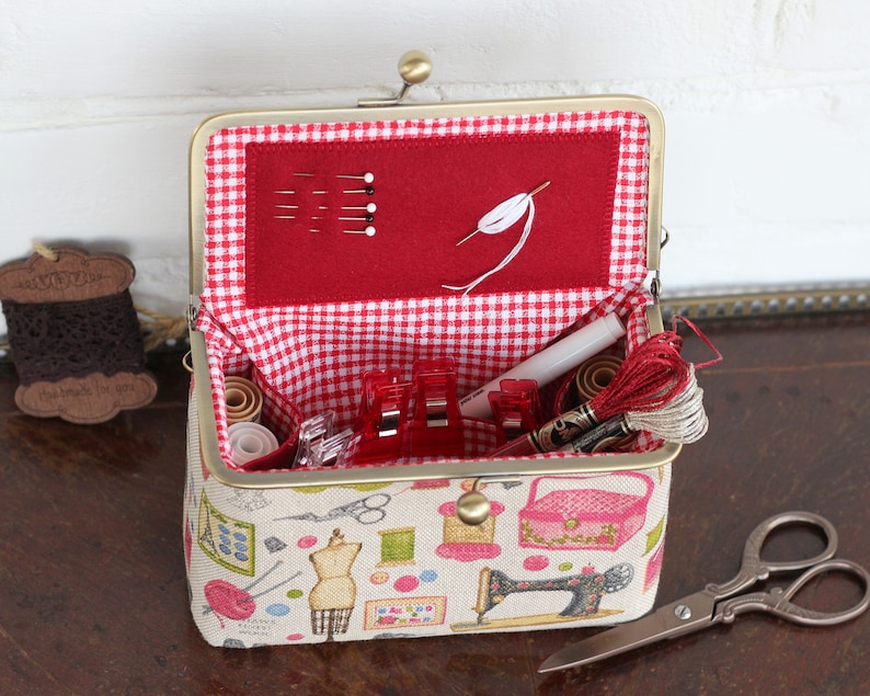 May include: A rectangular sewing organizer with a brass frame and clasp, featuring a beige fabric exterior with colorful sewing-themed illustrations. The interior is lined with red and white gingham fabric and contains sewing needles, thread, and clips.