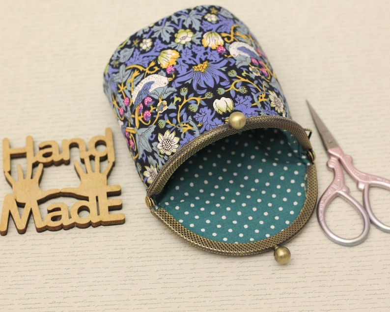 May include: A fabric pouch with a floral and bird pattern in shades of blue, yellow, and white, featuring a bronze-toned metal kiss-lock clasp and a teal interior lining with white polka dots, accompanied by wooden Hand Made text and scissors.