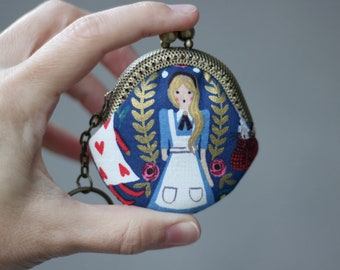 May include: A small, round coin purse with a brass clasp and chain. The purse features a blue fabric with an illustration of Alice in Wonderland, gold leaves, and red roses. The other side has a white background with red hearts.