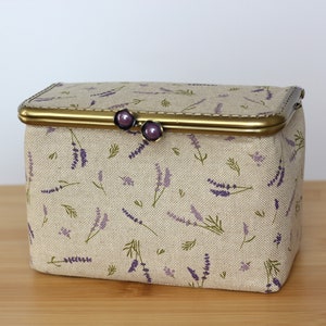 May include: A rectangular fabric storage box with a beige base and a lavender floral print. The box features a metallic brass-toned frame with a hinged closure topped with two small, round, dark purple decorative knobs for opening and closing.