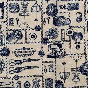 May include: A cream-colored fabric featuring a repeating dark blue print of sewing and crafting supplies, including scissors, spools of thread, buttons, dress forms, and a sewing machine, with a decorative label reading FEEL CHEERY.