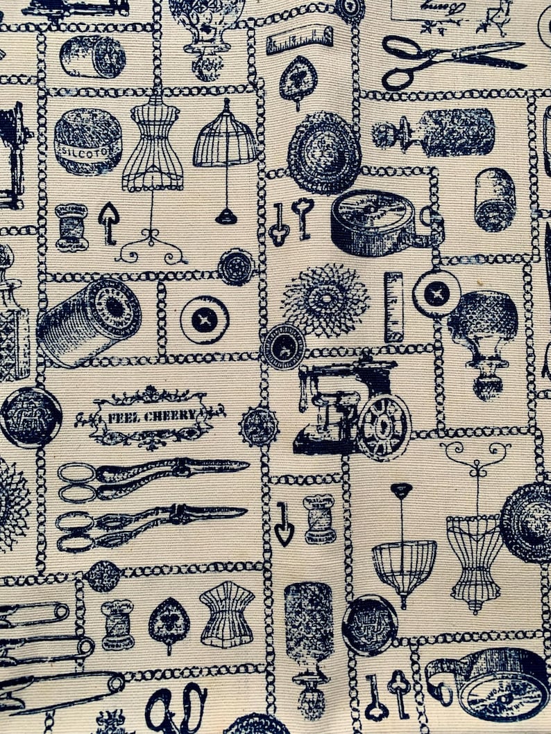 May include: A cream-colored fabric featuring a repeating dark blue print of sewing and crafting supplies, including scissors, spools of thread, buttons, dress forms, and a sewing machine, with a decorative label reading FEEL CHEERY.