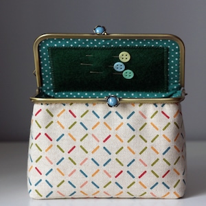 Puede incluir: Una pequeña bolsa de tela de color crema con un cierre de latón y detalles turquesa. La bolsa está forrada con tela verde y tiene un patrón de lunares blancos. La bolsa está abierta y tiene algunas agujas de coser y botones en su interior.