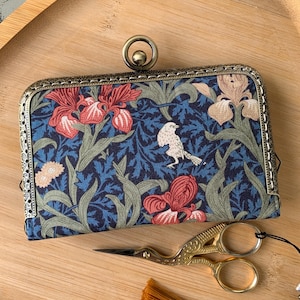 May include: A rectangular fabric coin purse featuring a dark blue background with a floral pattern of red and beige irises, green leaves, and a small white bird. The purse has an ornate antique brass metal frame with a circular clasp closure.