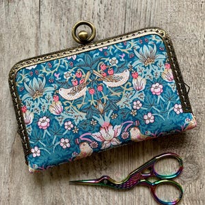 Può includere: Una pochette rettangolare in tessuto con sfondo verde acqua caratterizzata da un motivo ripetuto di uccelli, fiori rosa e fogliame verde. La pochette presenta una cornice in ottone in stile antico con una chiusura circolare. Accanto vi sono forbici iridescenti.