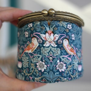 May include: A round, teal fabric coin purse with a brass clasp. The fabric features a floral and bird pattern in shades of teal, pink, and white. The purse is held in a hand.