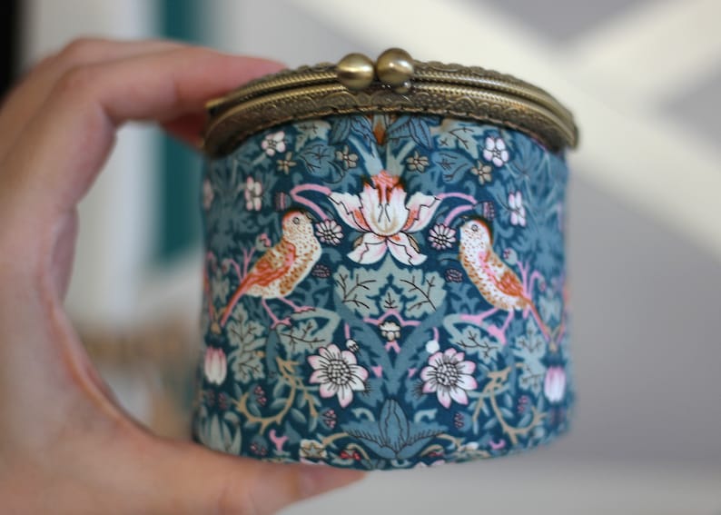 May include: A round, teal fabric coin purse with a brass clasp. The fabric features a floral and bird pattern in shades of teal, pink, and white. The purse is held in a hand.