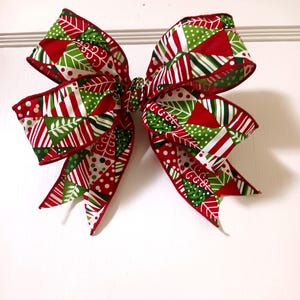 May include: A large decorative bow with a festive Christmas theme. The bow features a vibrant mix of red, green, and white patterns, including stripes, polka dots, and geometric shapes. The bow is likely made of fabric and is designed for holiday decorations.