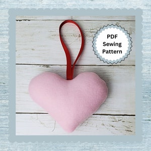 Può includere: Un cuore di feltro rosa con un nastro rosso appeso a uno sfondo di legno bianco. Il testo "PDF Sewing Pattern" è su un cerchio grigio con sfondo bianco.