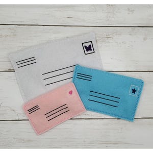 May include: Three felt envelopes in different sizes, with embroidered details. The largest envelope is white with a black butterfly, the medium envelope is light blue with a black star, and the smallest envelope is pink with a pink heart.