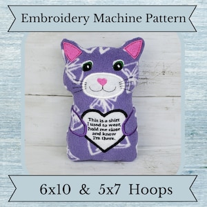 May include: A purple cat-shaped plushie with pink ears and a pink heart-shaped nose. The plushie has a white geometric pattern and a heart-shaped patch with text. The text reads: "This is a shirt I used to wear, hold me close and know I'm there."