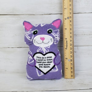 May include: A soft toy cat in purple and white, with embroidered details and whiskers. The doll features a heart-shaped design with the text: "This is a shirt I used to wear, hold me close and know I'm there."