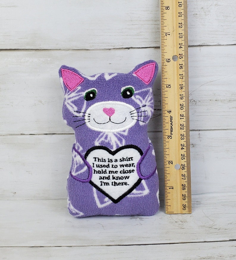 May include: A soft toy cat in purple and white, with embroidered details and whiskers. The doll features a heart-shaped design with the text: "This is a shirt I used to wear, hold me close and know I'm there."