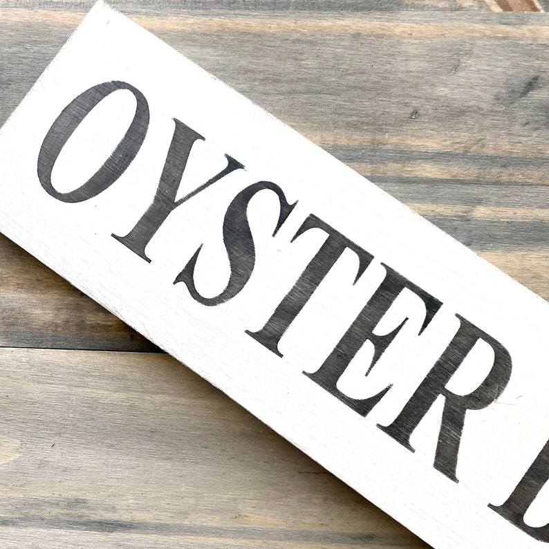 May include: A rectangular white wooden sign featuring the word OYSTER printed in large, bold, black serif capital letters. The sign is positioned at an angle, displaying a smooth painted surface with visible wood grain texture underneath the white paint.