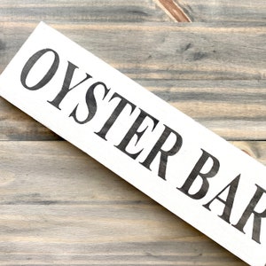 May include: A rectangular white wooden sign featuring the text OYSTER BAR in bold, black, capital letters. The sign is oriented diagonally across the frame, showcasing a clean, high-contrast design with visible wood grain texture on the surface.