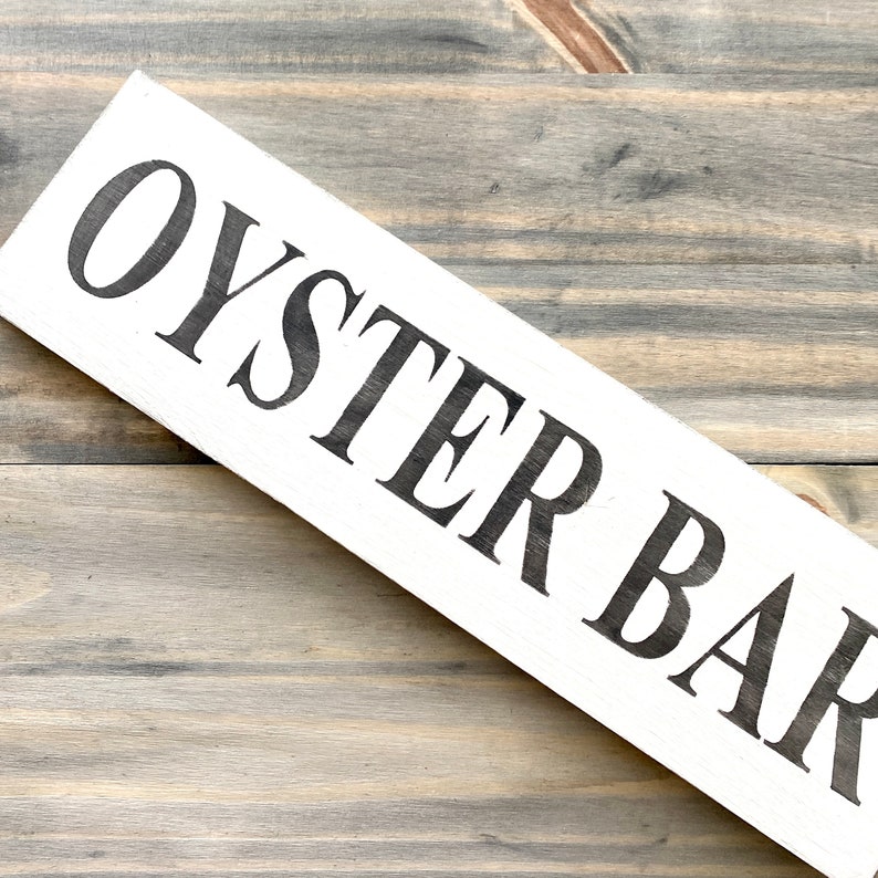 May include: A rectangular white wooden sign featuring the text OYSTER BAR in bold, black, capital letters. The sign is oriented diagonally across the frame, showcasing a clean, high-contrast design with visible wood grain texture on the surface.