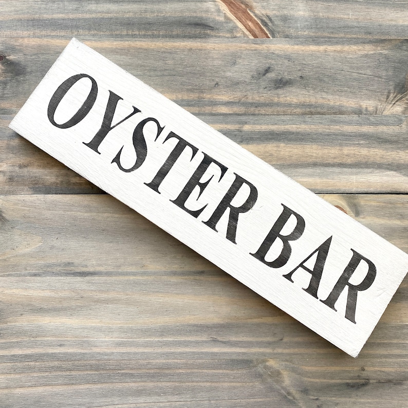 May include: A rectangular white wooden sign with the words OYSTER BAR printed in large black capital letters. The sign features a visible wood grain texture and is positioned diagonally across the frame, showcasing a clean and simple design.
