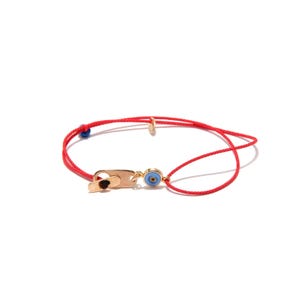 May include: A red string bracelet featuring a gold-toned metal clasp and a small circular charm with a blue and white evil eye design. The bracelet includes a tiny blue bead accent on the cord, providing a simple and adjustable design.