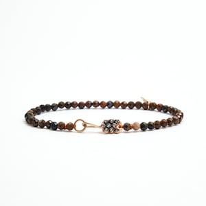 May include: A bracelet with faceted brown and dark blue beads. The bracelet has a rose gold clasp with a decorative cluster of small, dark beads. The bracelet is displayed against a white background.