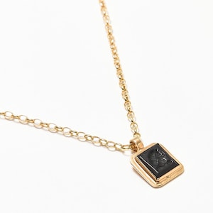 May include: A gold-colored necklace with a square pendant. The pendant features a black, carved design within a gold frame. The chain is also gold and has a delicate link design. The necklace is displayed on a white background.