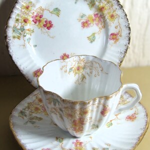May include: A vintage tea set comprising a teacup, saucer, and plate. The white porcelain has a floral design with pink and yellow flowers and green leaves. The edges are scalloped and finished with gold trim.